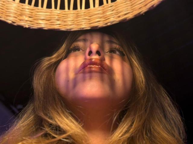 AngeliqueGirard's face with wavy blonde hair, lips slightly parted, looking upward. Shadows from a woven hat pattern across her face, giving a warm, intimate look.
