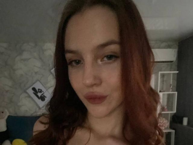 Een aantrekkelijke vrouw met rood haar, volle lippen en subtiele make-up, die de camera aankijkt met een lichte glimlach, in een kamer met weinig licht. Haar borsten zijn niet zichtbaar..