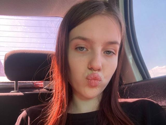 DignaHottie with puffy red lips making a kissing face in a car, sunlight highlighting her brown hair, wearing a black shirt, looking slightly annoyed.