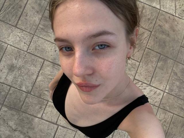 Il selfie di DignaHottie la mostra con gli occhi azzurri, lentiggini e un leggero sorriso, che indossa una maglietta nera, guarda verso l'alto verso la fotocamera, su un pavimento piastrellato..