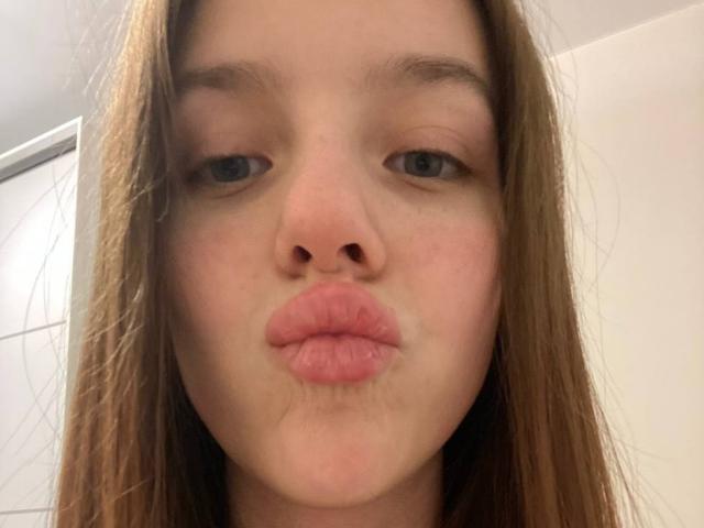 DignaHotties voluminöse, rosa Lippen sind zu einem albernen, küssenden Gesichtsausdruck geformt, mit blauen Augen, langen braunen Haaren und heller Haut auf einem Selfie..