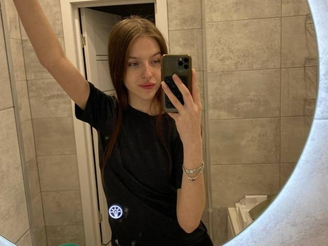 DignaHottie with straight brown hair, black t-shirt, and subtle pink lipstick, taking a mirror selfie with an arm raised, wearing a silver bracelet.