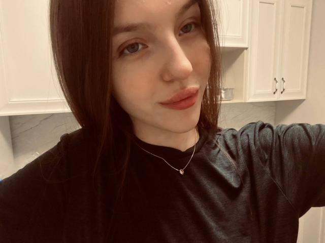 DignaHottie has pale skin, long brown hair, and is wearing a black shirt with a silver necklace. She's smiling with red lips and blue eyes in a selfie.