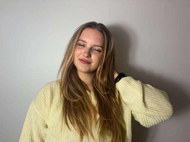 RosePlayful69 with long blonde hair, wearing a yellow knit sweater, smiling slightly, eyes closed, hand in hair, black wristband, soft light, casual, relaxed, cute.
