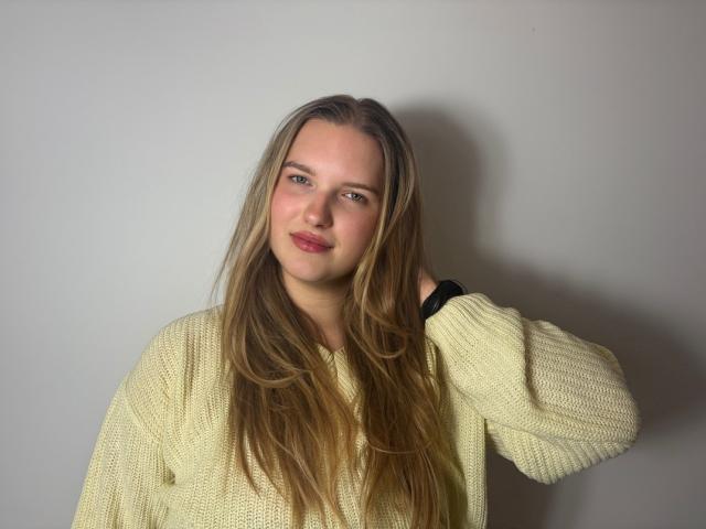 RosePlayful69 has long blonde hair, blue eyes, and is wearing a yellow knit sweater, standing with a slight smile and her right hand in her hair.
