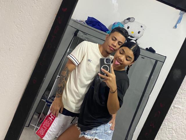 SweetVickyX with short brown hair, tattooed arm, white tee, and white shorts, standing next to SweetVickyX with black cap, black tee, and denim shorts, in a mirror selfie.