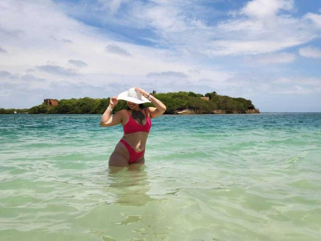 AlinaMatei in a red bikini stands in clear, shallow water, holding a white sunhat over her head, showing her curvy, tanned, bare-assed, and medium-sized tits.