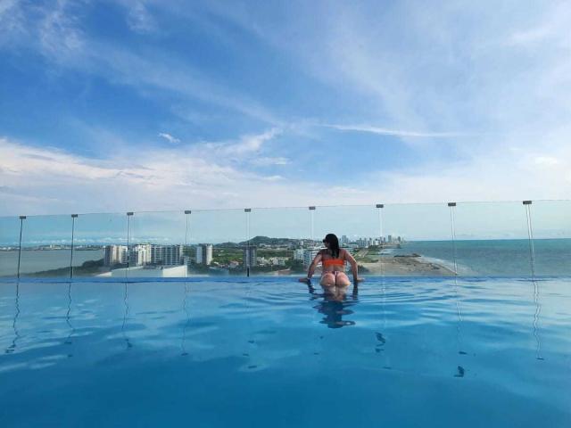 AlinaMatei with ass up in blue pool, wearing orange bikini, back to camera, glass fence, clear sky, ocean, city skyline in background, her butt and back are visible.