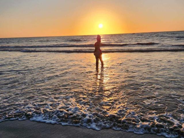 Silhouetted AlinaMatei stands in shallow ocean water, back to camera, sun setting, reflecting golden light on her ass and upper back, waves gently touching her legs.