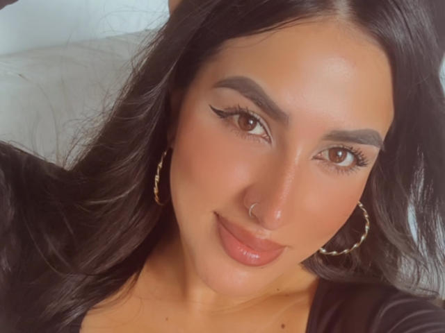 AlanaHarris has a cute face, brown eyes, and long black hair. She's wearing gold hoop earrings and a black top, with a small nose ring and glossy pink lips.