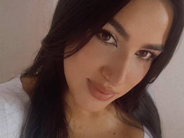 AlanaHarris has brown eyes, dark brown hair, and a small nose ring. She's smiling with her lips slightly parted, wearing a white top. Her ass is not visible.
