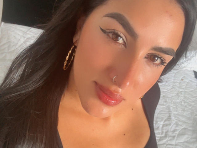 AlanaHarris has dark hair, brown eyes, bold eyebrows, winged eyeliner, red lipstick, hoop earrings, nose ring, and a slight smile, wearing a black top.