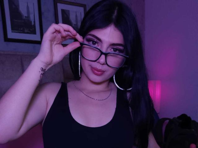 IsabellaaRossi with glasses, black tank top, hoop earrings, and a chain, touching her glasses, showing her tits, and a tattoo on her right arm. Purple light.
