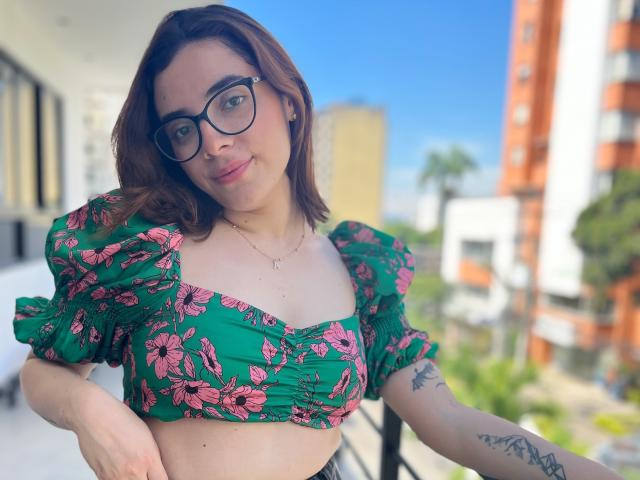 Lia, portant des lunettes, un haut court à fleurs vertes, des manches bouffantes, montrant ses seins, des cheveux bruns mi-longs, un tatouage sur l'avant-bras gauche, un sourire discret, debout sur un balcon, un ciel bleu clair..