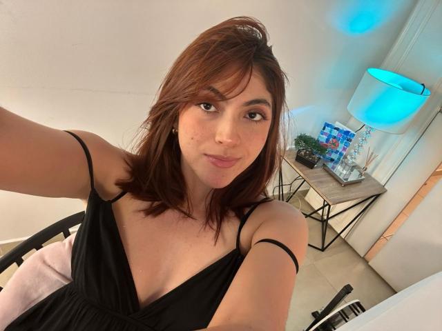SpencerLe with brown hair, wearing a black dress, showing small tits, cute face, and a slight smile, sitting in a room with a blue lamp and a wooden table.