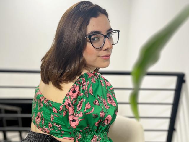 Lia, met bruin haar tot op de schouders, een bril, een groene jurk met bloemen, roze bloemen, lachend naar de camera, een witte muur, een zwarre metalen leuning, een groen blad op de voorgrond..
