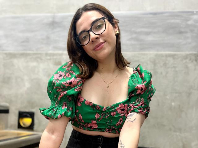Lia, con gafas, lleva un top corto de color verde con estampado floral, mostrando sus pechos, jeans negros y un tatuaje en su brazo derecho, sonriendo ligeramente, en un ambiente interior informal..