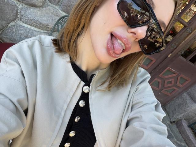 SensualMila with tongue out, wearing black sunglasses, gray jacket, and black top with silver buttons, has light brown hair, and a fair skin tone.