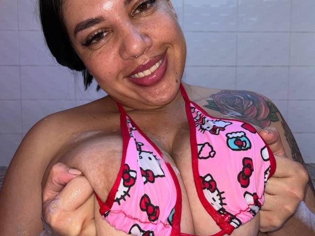 AndreaBusty with wet skin, smiling, holding up pink Hello Kitty bikini top, showing big tits, rose tattoo on right shoulder, nose ring, dark hair, and glossy pink lips.
