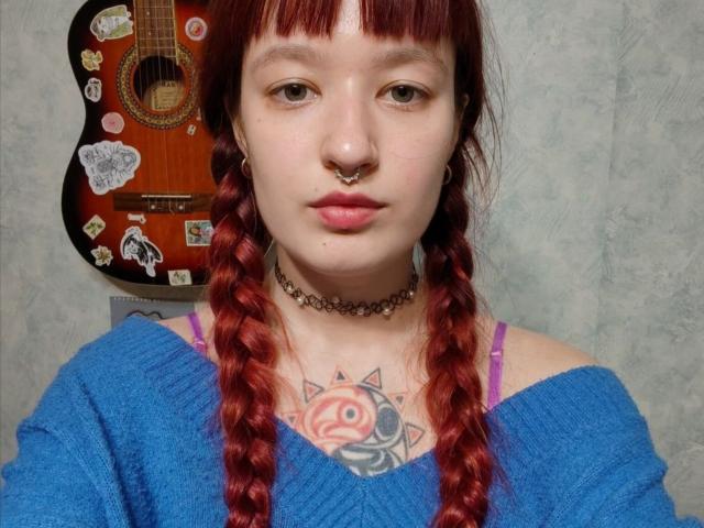 ElaneVuono mit roten Zöpfen, Nasenring und tätowierter Brust, trägt einen blauen Pullover und eine Halskette. Im Hintergrund ist ein Gitarre mit Aufklebern zu sehen, und man kann lila BH-Träger erkennen..