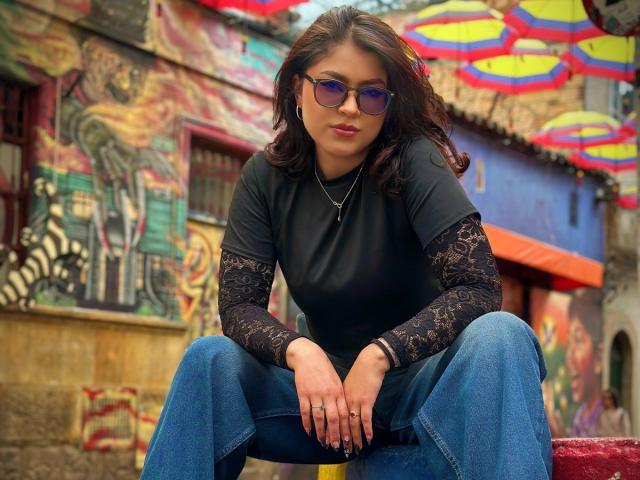 VioletteBlake with purple sunglasses, black lace sleeves, black top, blue jeans, sitting with legs apart, hands on knees, looking at camera, in colorful, graffiti-filled urban setting.