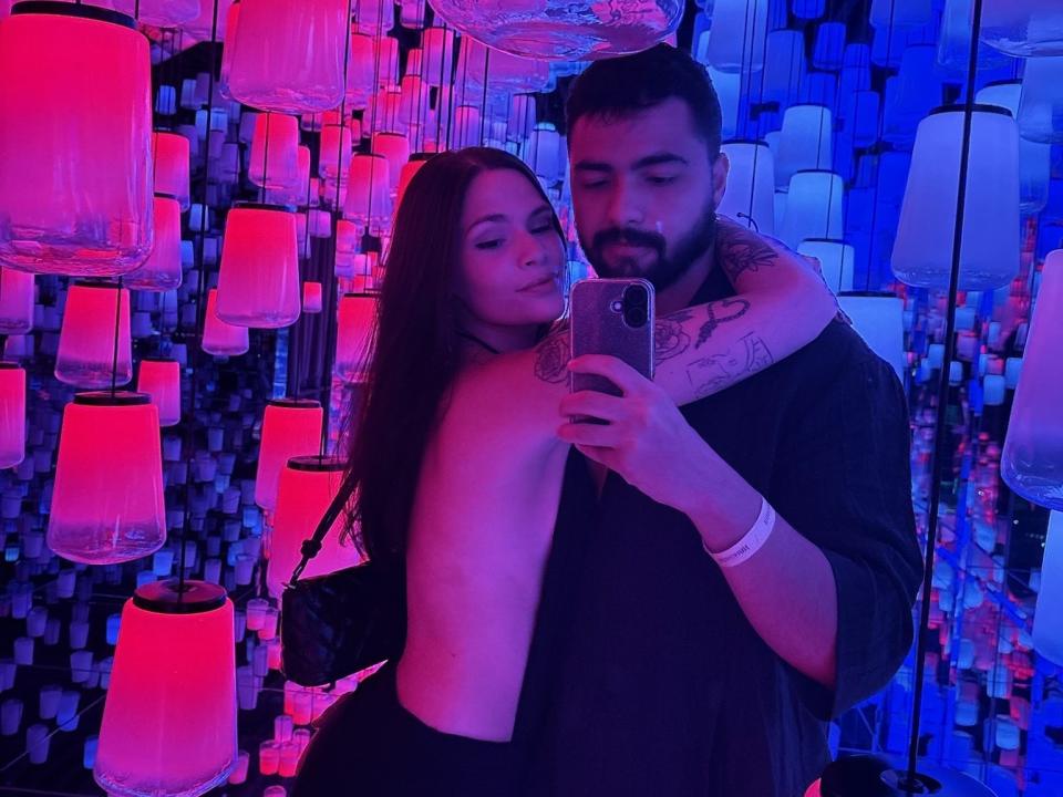 GiovannieLaura, onthuld tot haar borsten, leunt tegen een man met een baard en tatoeages, en maakt een selfie in een kamer met blauw en roze verlichting, met lantaarns die aan het plafond hangen. Hij draagt een zwart shirt..