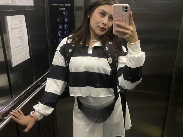 IsabellaPalacio with brown hair, wearing a black and white striped shirt, small floral patches, white pants, and a black pouch, taking a selfie in an elevator.