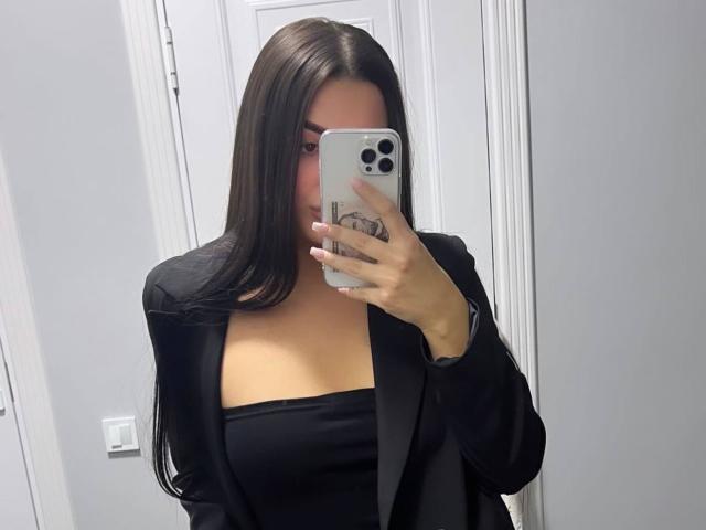 Dominique, met lang zwart haar, maakt een foto van zichzelf in de spiegel, gekleed in een zwart blazer over een zwarte top zonder bandjes, en houdt een iPhone met een witte hoes vast, waarbij haar borsten zichtbaar zijn..