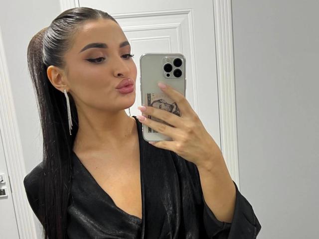 Dominique: Wow, met glad achterover gekamd zwart haar, een zwarte satijnen badjas aan, met een frons op haar lippen, een zilveren iPhone vasthoudend, een spiegel-selfie makend, waarbij ze haar nagels en subtiele make-up laat zien..