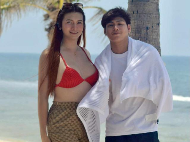 KateSteve with long brown hair, red bikini top, and checkered skirt, stands next to KateSteve with short black hair, white shirt, and towel, by the ocean.