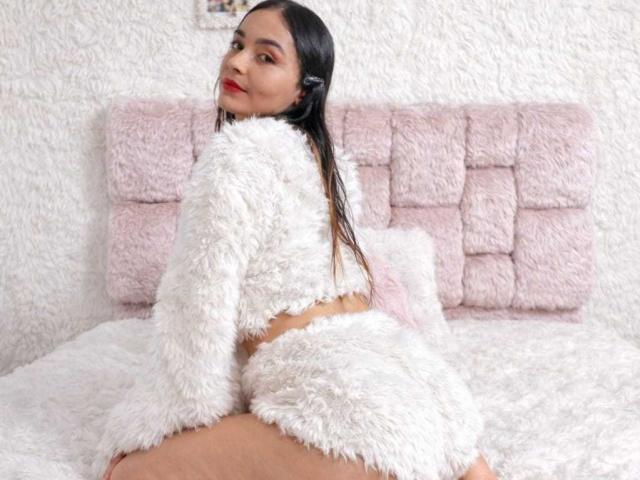 MindyLove with long black hair, wearing fluffy white lingerie, looking over her shoulder, sitting on a pink, fluffy bed, showing her ass and smiling with red lipstick.