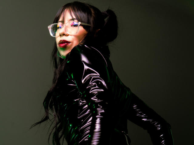 MiaDeLune with long black hair in pigtails, wearing clear glasses, shiny black latex dress, and green light reflecting off her outfit, looking over her shoulder with red lipstick.