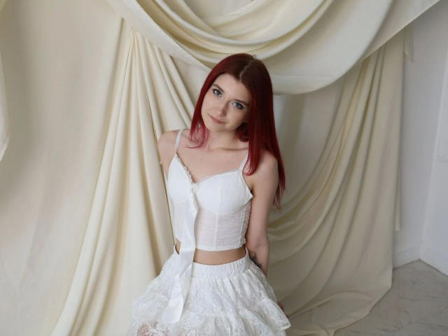Monnyca-hot with red hair, wearing a white lace bustier and skirt, sits against draped fabric. She has small tits and a cute face, looking slightly to the side.