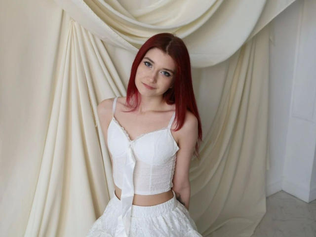 Monnyca-hot with red hair, small tits, wearing a white lace-up bustier and skirt, standing against draped fabric, looking at camera with a slight smile.