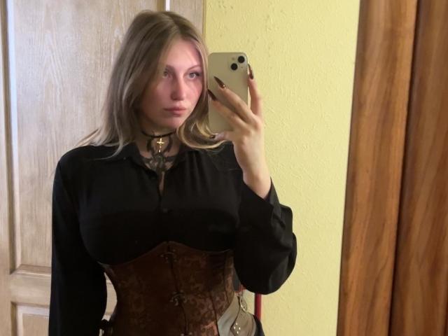MelinaBoye with blonde hair, black shirt, brown corset, and choker, taking a mirror selfie with her phone, showing her tits and holding her phone with painted nails.