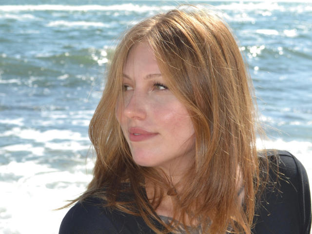MelinaBoye with light brown hair, slightly wavy, and a subtle smile, wearing a black shirt, standing by the ocean with gentle waves in the background.