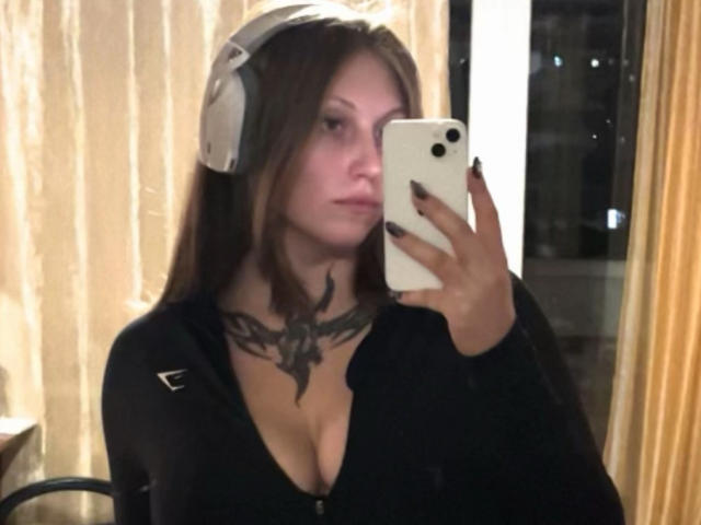 MelinaBoye with headphones, black zip-up top, cleavage, black nail polish, holding phone, tattoo on chest, mirror selfie.
