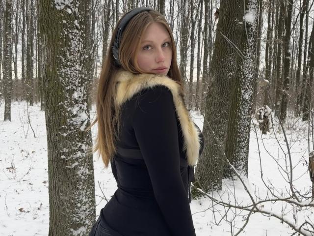 MelinaBoye with long brown hair, black headphones, black long-sleeve top with yellow fur trim, standing in snowy forest, looking back, showing her ass.