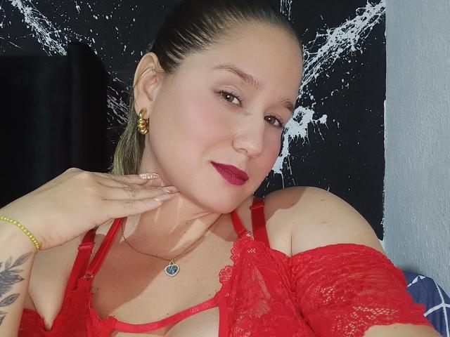 AlexaHotSeins with red lace bra, red lipstick, and gold earrings, hand on chin, brown eyes, tattoo on wrist, necklace with green gem, dark hair pulled back.