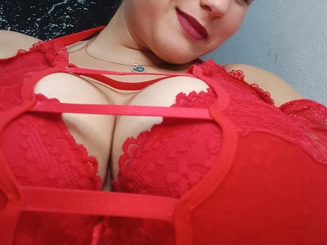 AlexaHotSeins in red lace lingerie, showing her big tits, with a red strappy top, and a necklace, smiling with red lipstick, in a slightly angled selfie.