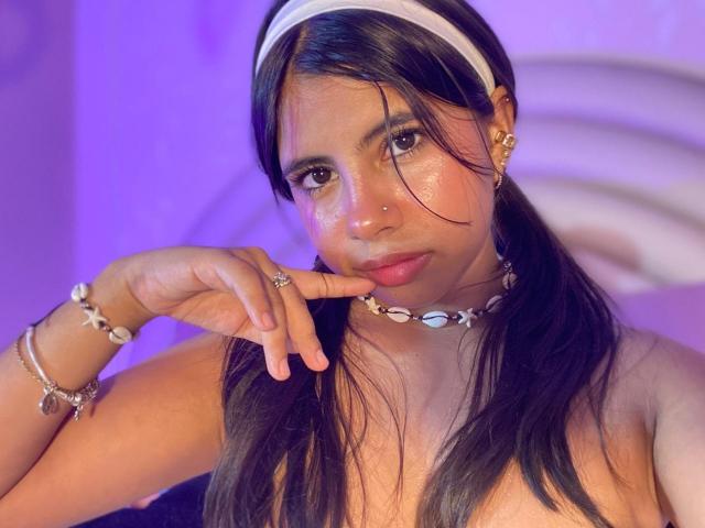 CelesteMorel with long black hair, white headband, shell necklace, and earrings, touching her lip with a finger, wearing a silver bracelet, in a purple-lit room.