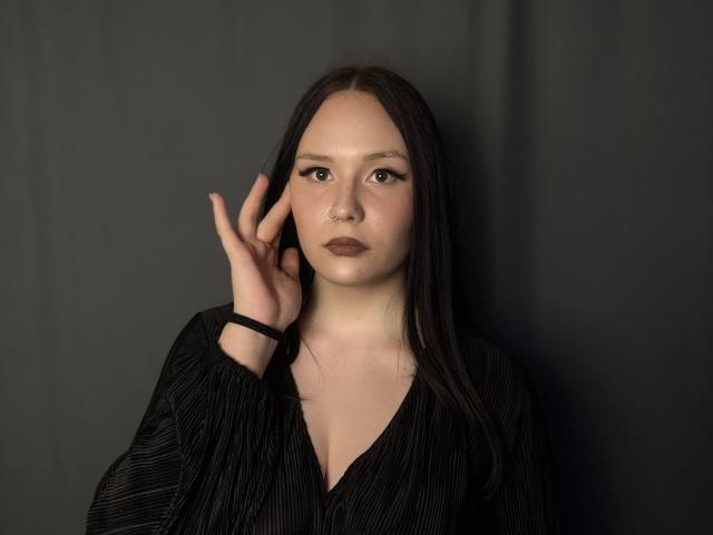KortneyKempf hat langes, schwarzes Haar, einen Nasenring und trägt ein schwarzes Hemd mit Streifen. Sie hebt ihre rechte Hand gegen einen dunklen Hintergrund. Ihr Gesichtsausdruck ist ernst..