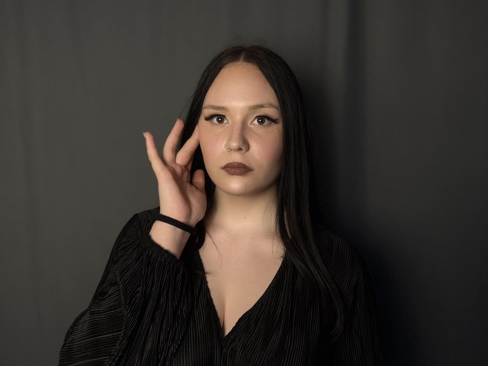 KortneyKempf has long black hair, a nose ring, and is wearing a black pinstriped shirt, raising her right hand against a dark background. Her face is serious.