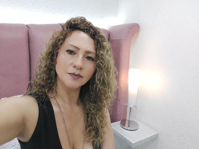 AlexaFireHot, con i capelli biondi ricci, indossa una canotta nera e ha un poggiatesta rosa intenso, mostrando il seno e un trucco leggero in una camera da letto luminosa. Una lampada è posizionata su un comodino bianco..