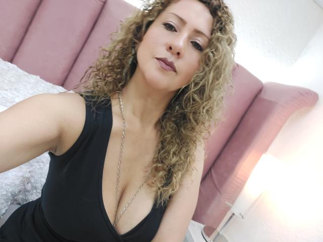 AlexaFireHot mit lockigem, blondem Haar, trägt ein schwarzes, tief ausgeschnittenes Kleid, das ihre Brüste zeigt, und eine silberne Halskette. Sie blickt mit einem leichten Lächeln in die Kamera..