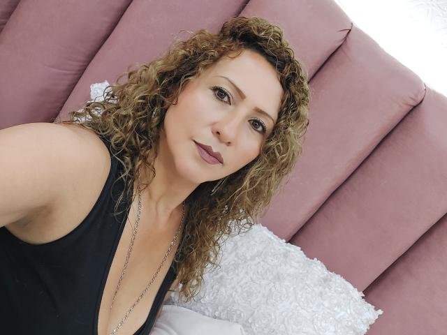 AlexaFireHot tiene el pelo castaño rizado, un tono de piel claro y lleva puesto un top negro con un escote pronunciado en V, que deja ver su escote. Está mirando a la cámara con una ligera sonrisa..
