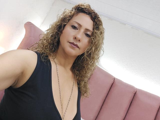 AlexaFireHot, con i capelli biondi ricci, indossa una maglia nera scollata che mette in mostra il seno, e una collana, guardando la fotocamera con un sorriso appena accennato..