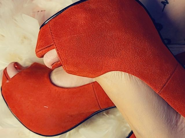 ElixirShanna's foot in a red, suede, high-heeled shoe, with her pink, trimmed toe nail visible, against a fluffy, white, fabric background.