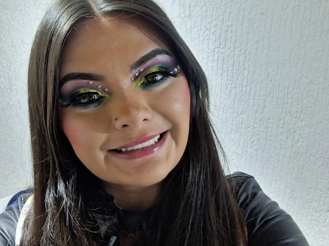 KatherinLatin with green and purple eye makeup, black eyeliner, and small facial piercings, smiling with straight brown hair and wearing a black shirt, showing her shiny white teeth.