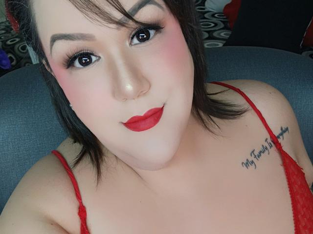 GiiannaMorrys has red lipstick, pink blush, black eyeliner, and red lace lingerie. She's smiling with a nose ring, dark brown hair, and a tattoo on her right shoulder.
